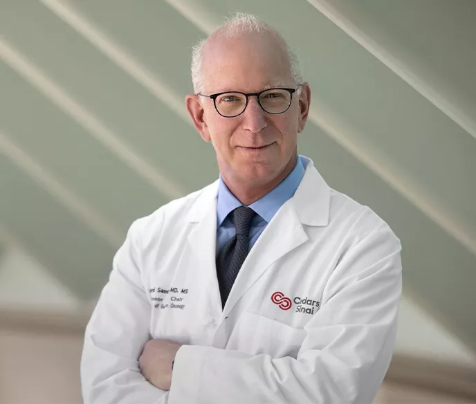 Older white male with glasses wearing a doctor's jacket that says "Cedars-Sinai" standing with his arms crossed