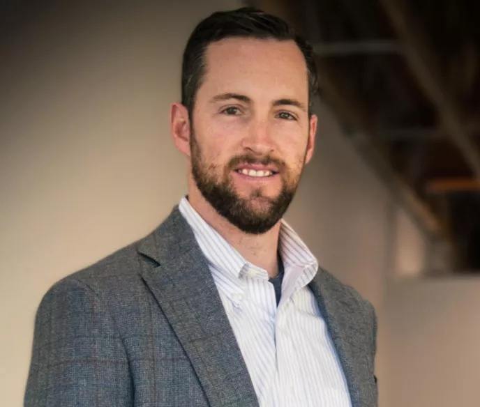 Dr. Ryan Owen headshot, white man with brown hair and beard standing in a light shirt and gray jacket