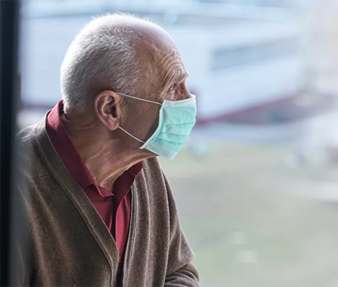 An old white man wearing a mask looking out a window