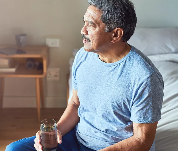 A Hispanic man sitting on the edge of his bed with a glass of water