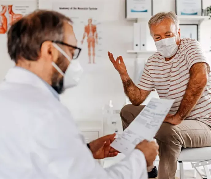 A man sitting on a hospital table wearing a mask while his doctor, also wearing a mask, is reading a paper