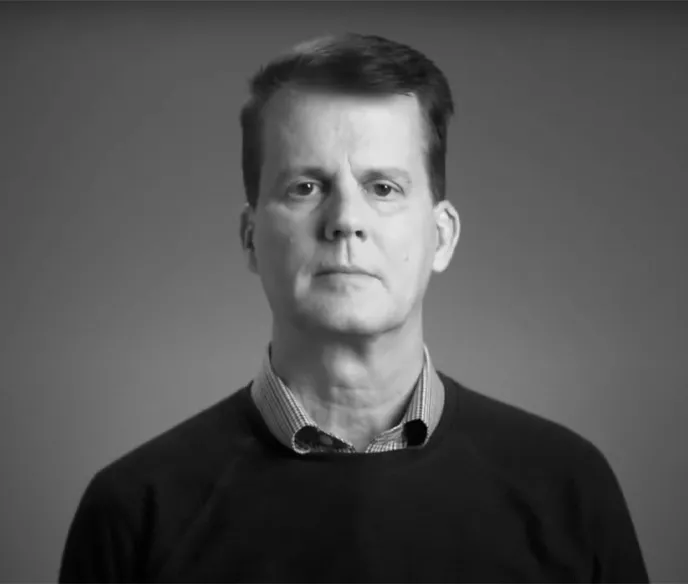 A black and white image of a white man wearing a dark sweater with a collared shirt underneath