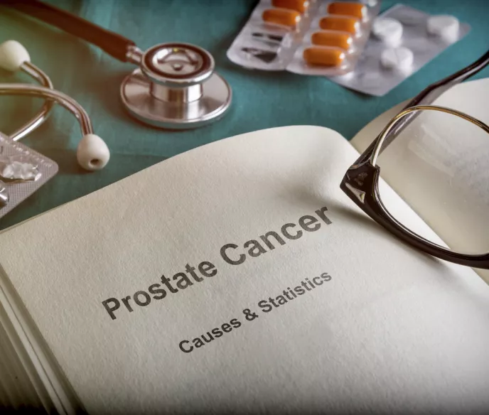 A book opened to a page with text that says "Prostate Cancer Causes & Statistics". There are glasses on the book and prescription pills on the desk.