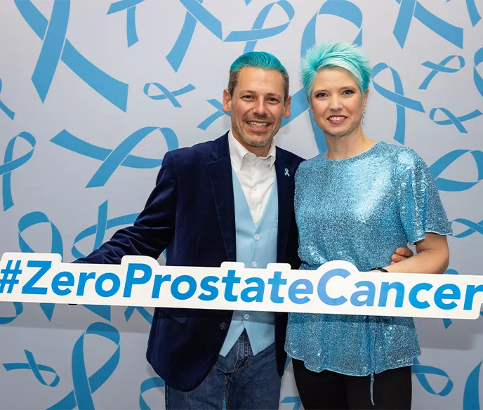 A couple with blue hair holding a sign that says #ZEROProstateCancer in front of a backdrop full of blue ribbons