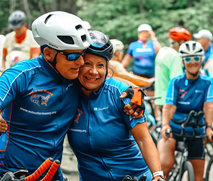 An older couple in cycling jerseys and helmets embracing while surround by many onlooking cyclists