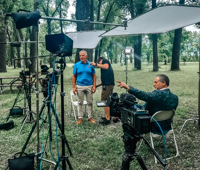 An older man in khaki pants and a blue shirt standing with lights and media all around him