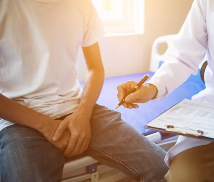 A shot of a man wearing a white tshirt and jeans sitting on a doctors table with his hands in his lap and a doctor sitting by the table on a stool holding a clipboard