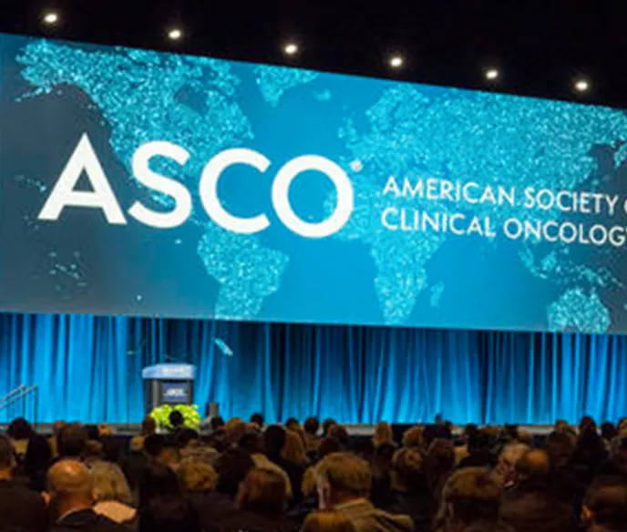 A large conference hall with a wide stage featuring a prominent display screen showing the ASCO (American Society of Clinical Oncology) logo and text in white against a turquoise world map background. The stage is framed by blue-lit curtains and lighting. The audience area is filled with attendees seated in rows, viewed from behind. The overall lighting creates a professional, dramatic atmosphere typical of major medical conferences.