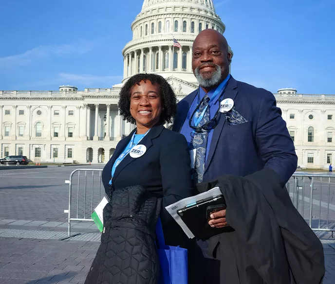 Two advocates wearing ZERO badges stand smiling in front of the U.S. Capitol building in Washington, D.C. on a sunny day. They are dressed professionally, with one person holding what appears to be documents or materials.