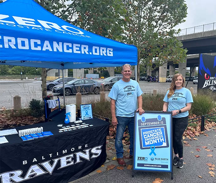 An outdoor awareness booth with a blue ZEROCANCER.ORG tent canopy. Two volunteers wearing light blue t-shirts stand by a table with Baltimore Ravens branding and prostate cancer awareness materials. One volunteer holds a sign announcing "September is Prostate Cancer Awareness Month" with a blue ribbon logo.