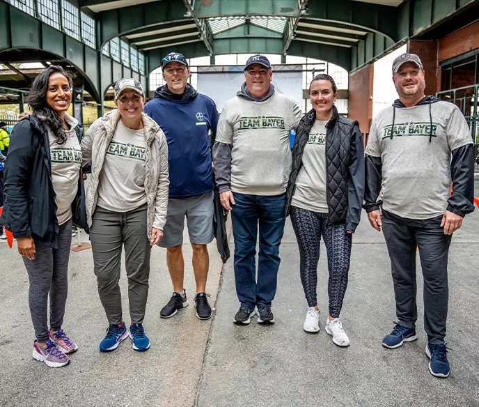 A group of six people wearing "Team Bayer" branded shirts and athletic wear stand together under what appears to be a covered outdoor venue. They are dressed casually and appear to be at a community event.