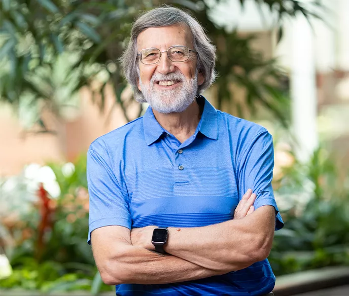 A man with gray hair and beard wearing a blue striped polo shirt smiles warmly at the camera with his arms crossed. He's wearing a smart watch and is photographed in front of a blurred green plant background.