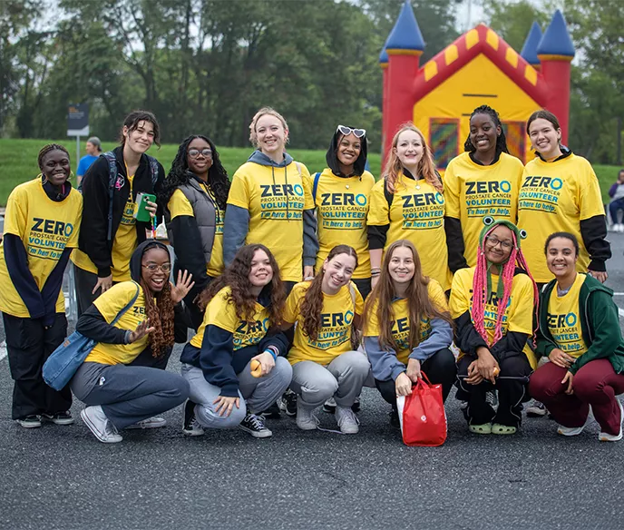 A group of enthusiastic volunteers wearing bright yellow ZERO t-shirts pose together outdoors, with some standing and others kneeling in front. A colorful bounce house castle is visible in the background. The diverse group shows genuine smiles and team spirit.