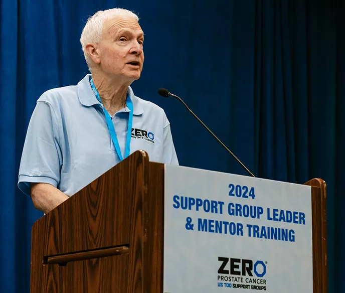 A speaker in a light blue ZERO polo shirt stands at a wooden podium with a sign reading "2024 Support Group Leader & Mentor Training" and the ZERO Prostate Cancer logo. Blue curtains form the backdrop.