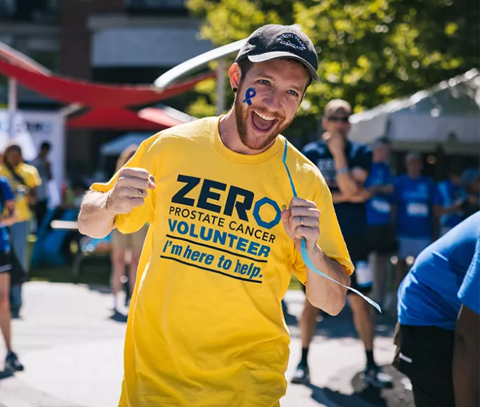 An energetic volunteer wearing a yellow ZERO Prostate Cancer Volunteer t-shirt with "I'm here to help" text smiles broadly at the camera on a sunny day. He has a blue ribbon painted on his face, with other volunteers visible in the background.