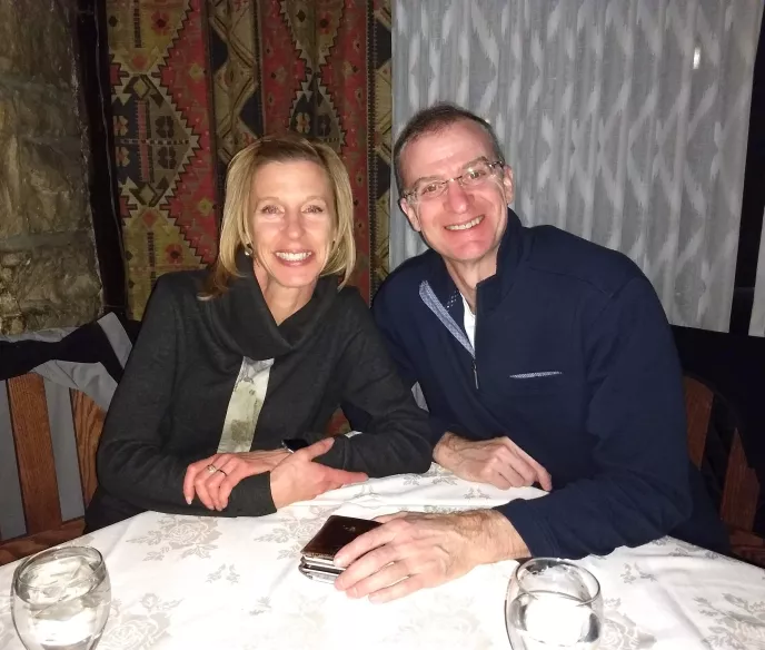 A couple sitting at a restaurant table with a decorative patterned tapestry on the stone wall behind them. The woman wears a black jacket and has blonde shoulder-length hair, while the man wears a navy blue quarter-zip pullover and glasses. Both are smiling warmly at the camera. The table has a white tablecloth with a floral pattern and two water glasses.