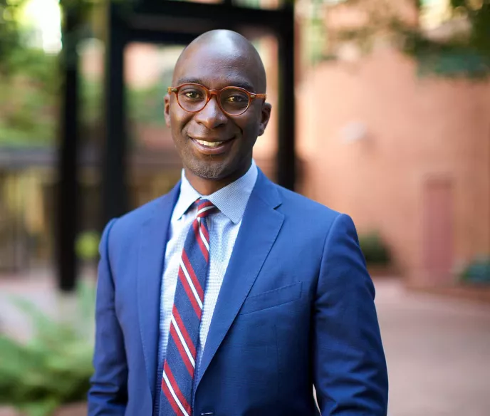 A professional headshot of a person wearing a blue suit, red and blue striped tie, and round brown-framed glasses. They are smiling warmly at the camera against a blurred outdoor background with greenery and brick walls. The photo is well-lit and captures a friendly, approachable demeanor.