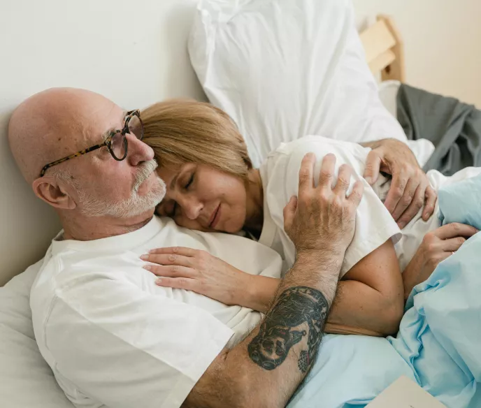 An elderly couple is lying in bed, cuddling closely. The man, wearing glasses and a white shirt, has a tattoo on his arm. The woman rests her head on his chest, eyes closed, with a content expression. They are covered with a light blue blanket.