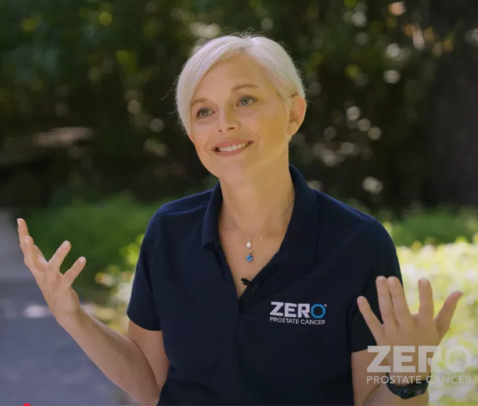 A woman with short, light blonde hair and a warm smile gestures expressively. She wears a dark blue polo shirt with the "ZERO Prostate Cancer" logo and a blue pendant necklace. The background features greenery, suggesting an outdoor setting.