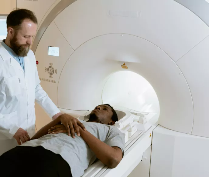 A patient lies on an MRI table, wearing a light gray t-shirt and dark pants, looking calm. A medical professional in a white lab coat stands beside them, focused and attentive. The MRI machine's circular opening and control panels are visible, creating a clinical and calm atmosphere.
