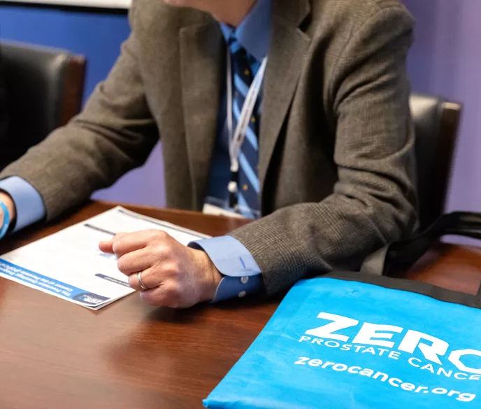 A person in a suit gestures while discussing materials on a table. A blue tote bag with "ZERO PROSTATE CANCER" is prominently displayed, highlighting prostate cancer awareness.