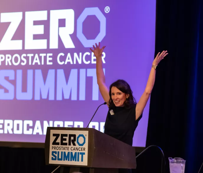 A woman with medium-length brown hair, wearing a black dress, stands at a podium with her arms raised enthusiastically. The backdrop reads "ZERO Prostate Cancer Summit.