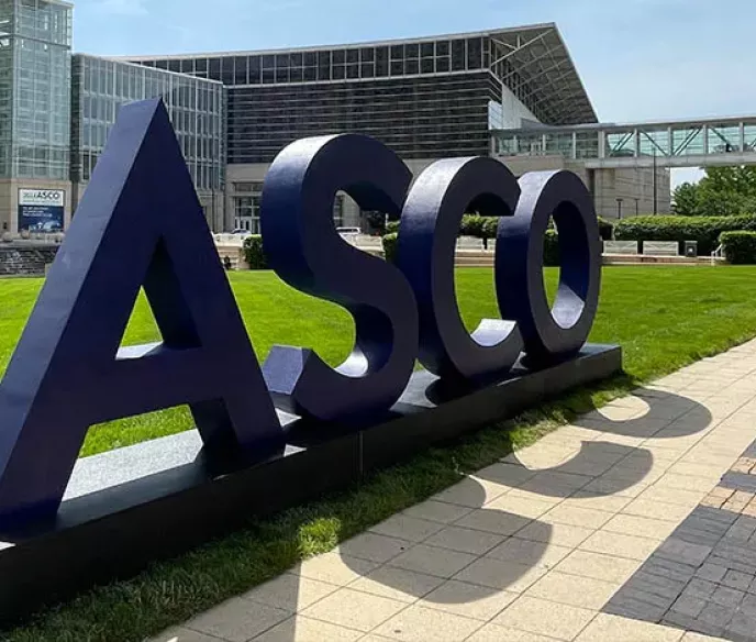 The image features large, bold letters spelling "ASCO" set against a green lawn. In the background, modern buildings with glass facades can be seen, along with walkways and trees, suggesting a bright and open outdoor environment.