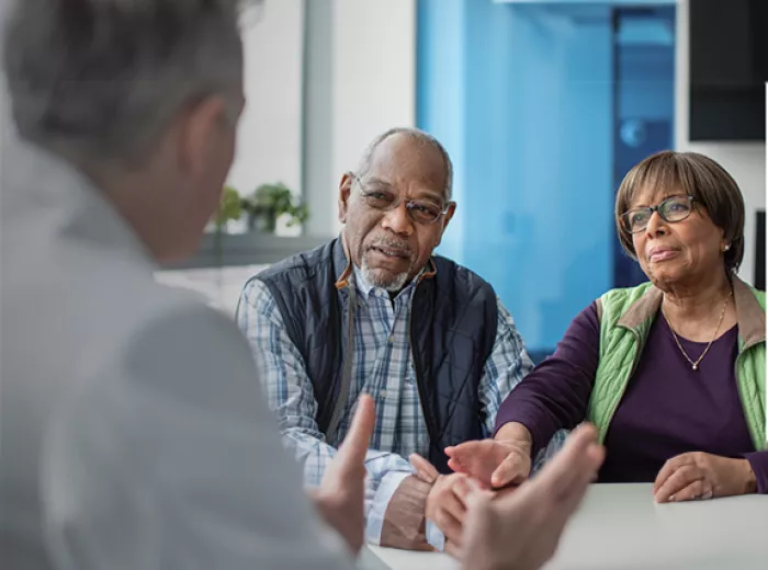 An older black couple sitting at a table in front of a doctor. They look to be engaging in conversation.