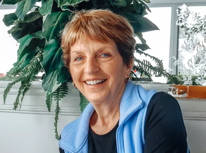 A woman with short reddish hair smiling while sitting in front of a window that has a large plant