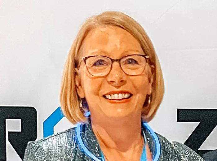 An older blonde woman with glasses standing against a ZERO sign