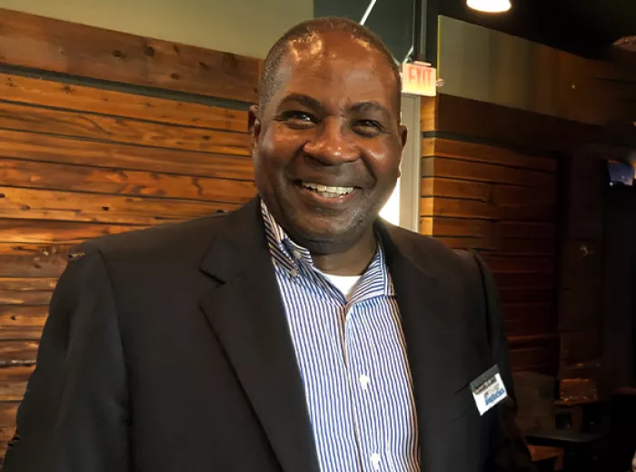 Black man in suit smiling in front of wood-paneled wall