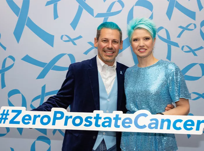 A couple with blue hair holding a sign that says #ZEROProstateCancer in front of a backdrop full of blue ribbons