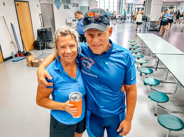 A couple, Scott and Katy Freitag, wearing cycling gear while standing together for a photo inside of a cafeteria