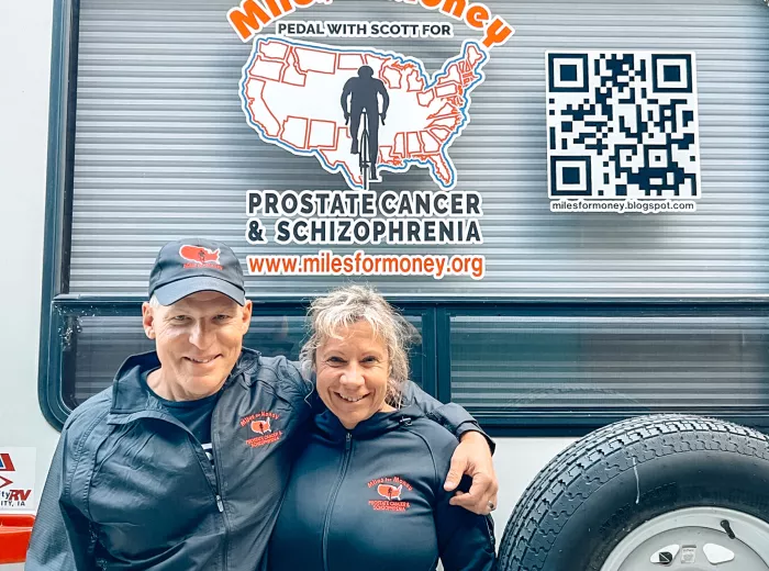 A couple, Scott and Katy Freitag, wearing black raincoats that have the Miles for Money logo standing outside their camper