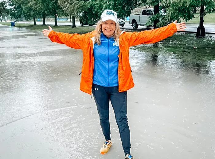 A woman in sporting gear, blue shirt, black leggings, orange raincoat, and a white ZERO Champion hat stands with her arms spread out as she embraces the rain. You can see a little flooding in the area she is standing in. 