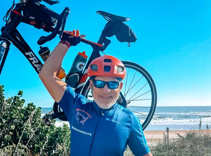 A man, Scott Freitag, wearing a blue cycling jersey and red helmet, holding a bicycle over his head