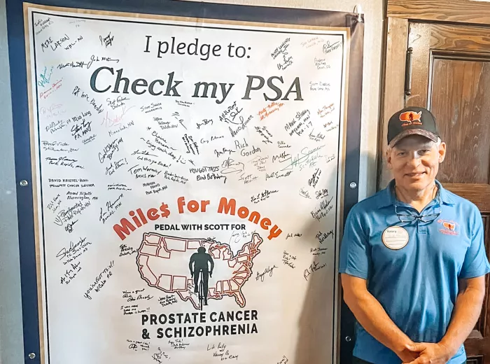 A man, Scott Freitag, wearing a blue polo shirt standing next to a banner that says "I pledge to Check my PSA" with the Miles for Money logo and has hundreds of signatures all over it