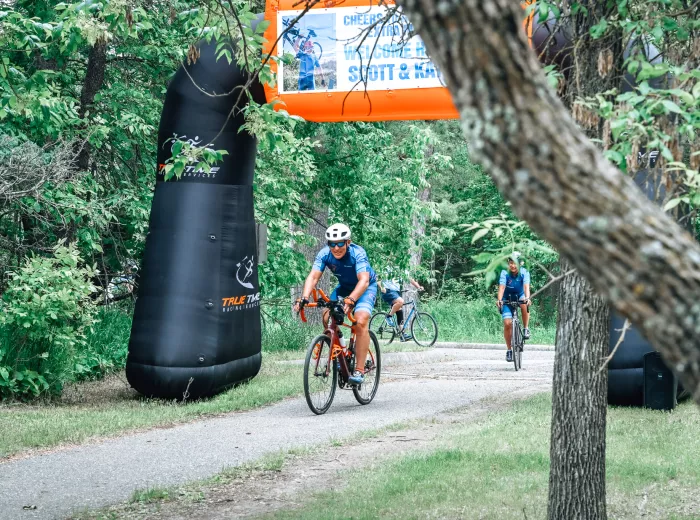 A man, Scott Freitag, several yards away riding in on his bicycle under a banner that says "Congratulations, Scott and Katy"