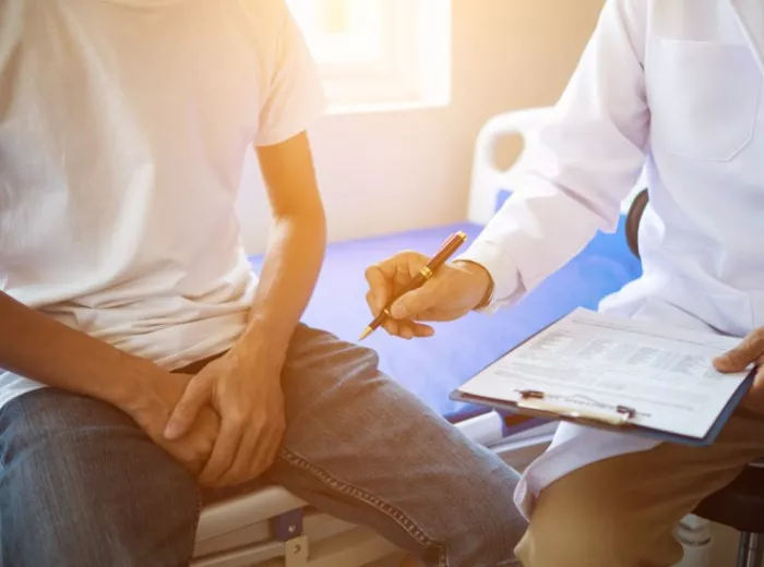 A shot of a man wearing a white tshirt and jeans sitting on a doctors table with his hands in his lap and a doctor sitting by the table on a stool holding a clipboard