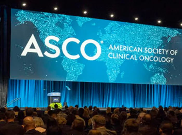 A large conference hall with a wide stage featuring a prominent display screen showing the ASCO (American Society of Clinical Oncology) logo and text in white against a turquoise world map background. The stage is framed by blue-lit curtains and lighting. The audience area is filled with attendees seated in rows, viewed from behind. The overall lighting creates a professional, dramatic atmosphere typical of major medical conferences.