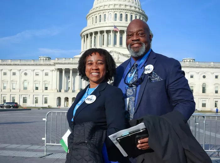 Two advocates wearing ZERO badges stand smiling in front of the U.S. Capitol building in Washington, D.C. on a sunny day. They are dressed professionally, with one person holding what appears to be documents or materials.