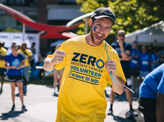 An energetic volunteer wearing a yellow ZERO Prostate Cancer Volunteer t-shirt with "I'm here to help" text smiles broadly at the camera on a sunny day. He has a blue ribbon painted on his face, with other volunteers visible in the background.