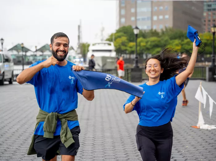 A man and a woman running joyfully, holding a blue Pfizer banner. They are wearing blue shirts, and the background shows a waterfront area with buildings and trees.