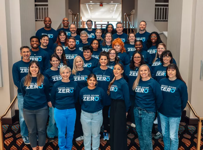 A large group of people standing on a staircase, all wearing matching navy blue shirts with the text "Bigger, Bolder, Zero" printed on them. They are smiling and posing for the photo in a well-lit indoor setting.