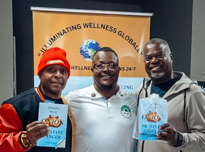Three men stand smiling in front of a banner that reads "Illuminating Wellness Globally." The man in the center wears a white polo shirt with a green wellness logo, while the two men on either side of him hold up copies of the book “Peeling Back the Onion on Prostate Cancer.” The man on the left wears a red hat and black-and-red varsity jacket, and the man on the right wears a light gray hoodie and sweatpants.