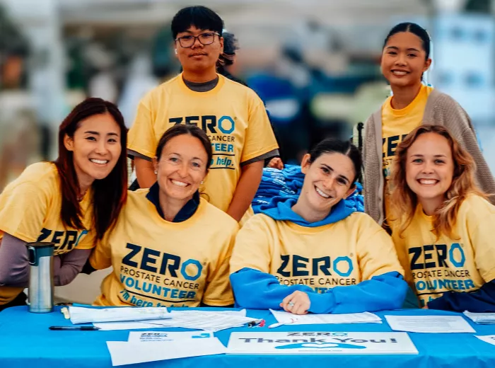 A group of six smiling volunteers, all wearing bright yellow “ZERO Prostate Cancer Volunteer” shirts, gather behind a registration table covered in papers and a blue tablecloth. They are outdoors at what appears to be a community event or race, with a blurred background of tents and people.