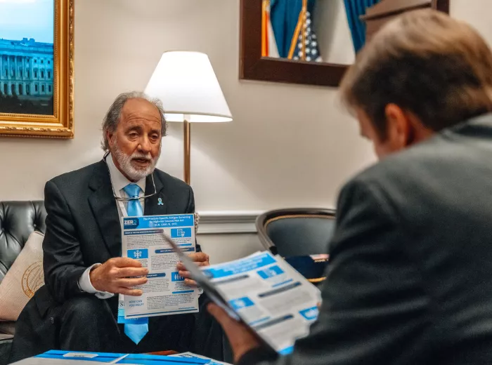 An older man with a white beard, wearing a dark suit and light blue tie, sits in an office setting holding a brochure related to prostate cancer legislation. He appears to be speaking to someone in the foreground whose back is to the camera. The room features official decor including U.S. flags and a framed photo of the Capitol.