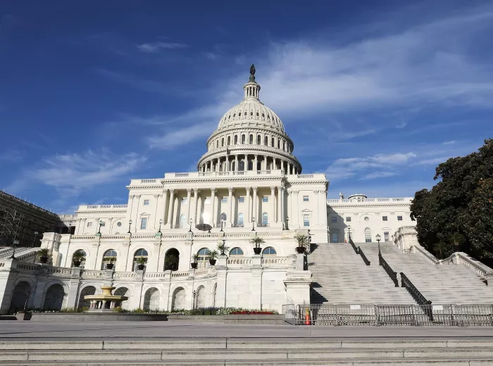 A grand building with a large dome topped by a statue, framed by a bright blue sky with wispy clouds. It features white columns and steps leading up to the entrance, with greenery and a fountain in the foreground.