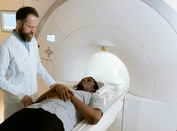 A patient lies on an MRI table, wearing a light gray t-shirt and dark pants, looking calm. A medical professional in a white lab coat stands beside them, focused and attentive. The MRI machine's circular opening and control panels are visible, creating a clinical and calm atmosphere.