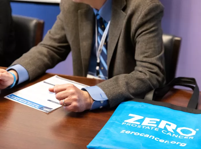 A person in a suit gestures while discussing materials on a table. A blue tote bag with "ZERO PROSTATE CANCER" is prominently displayed, highlighting prostate cancer awareness.